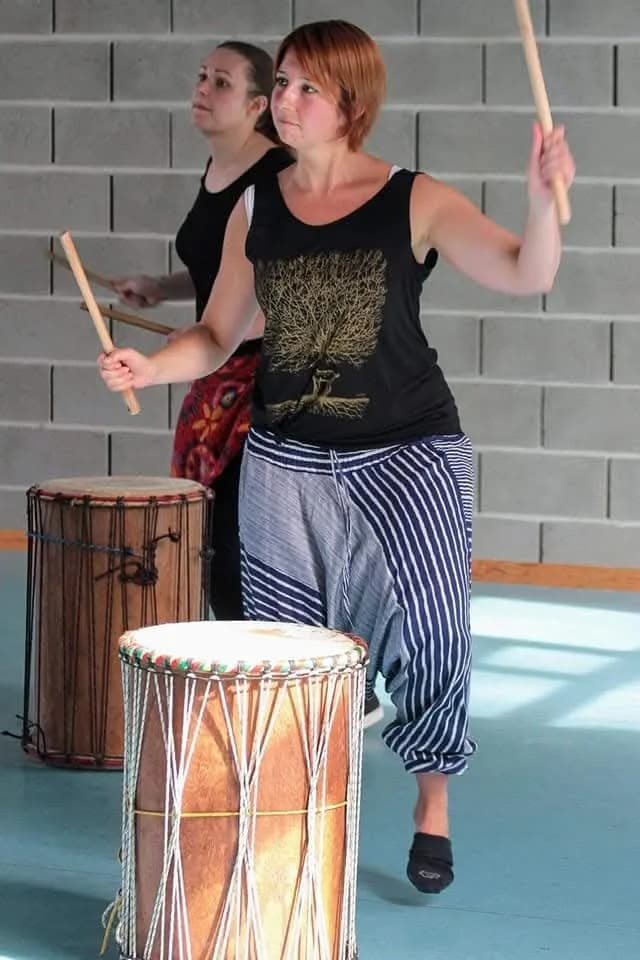 Une participante en pantalon rayé joue du djembé et du dundun aux baguettes, debout derrière ses instruments.