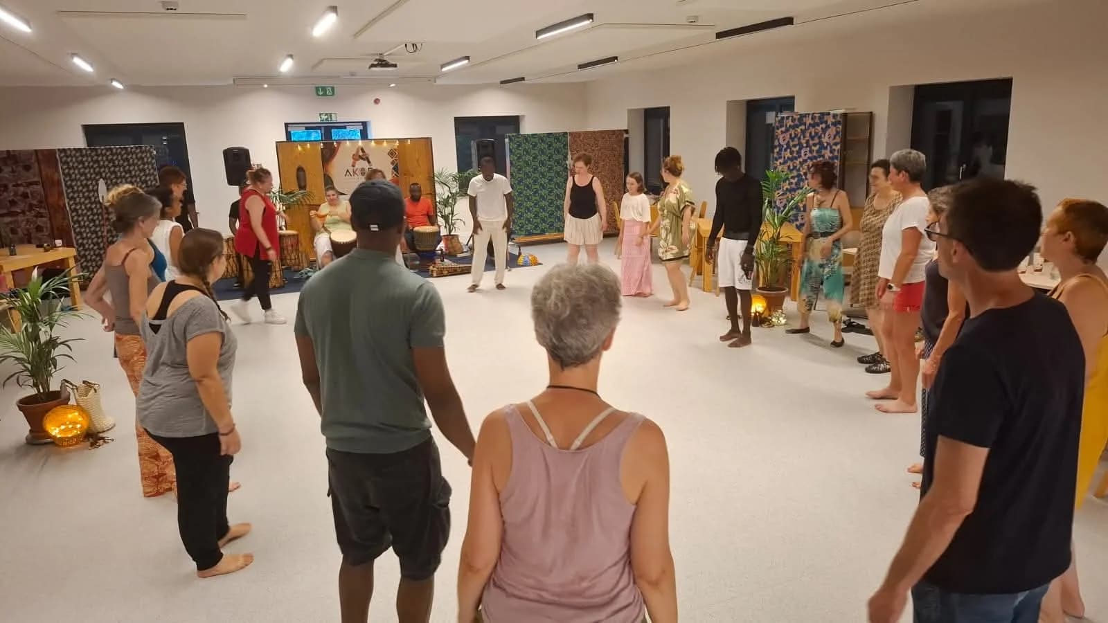 Un large cercle de participant·es debout dans la salle, entouré de tentures colorées et d'instruments.