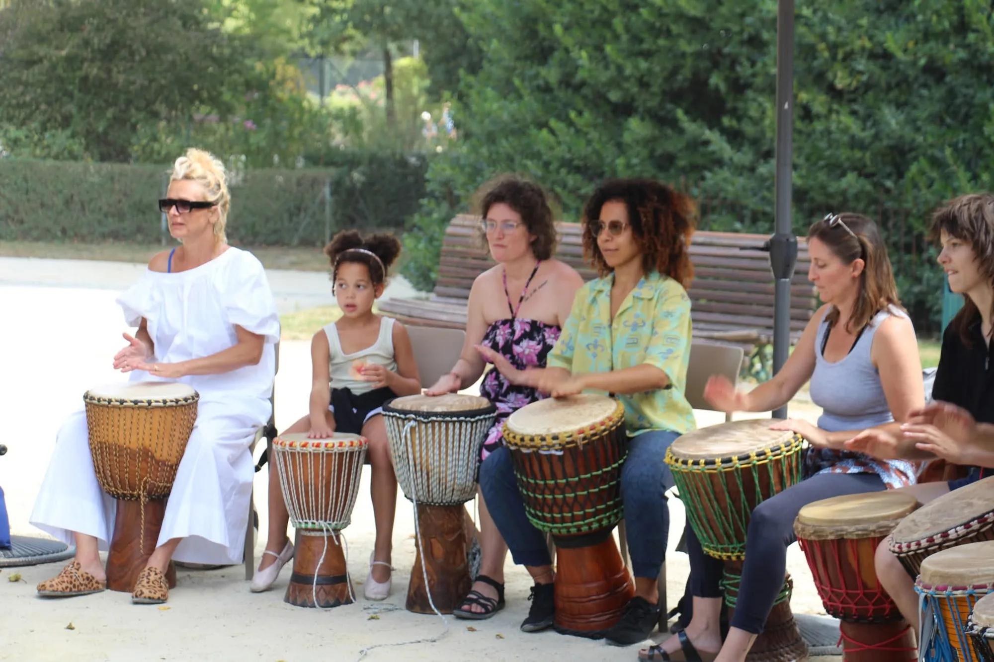 Percussions en plein air sous les arbres : plusieurs participantes et une enfant jouent du djembé, assises côte à côte.